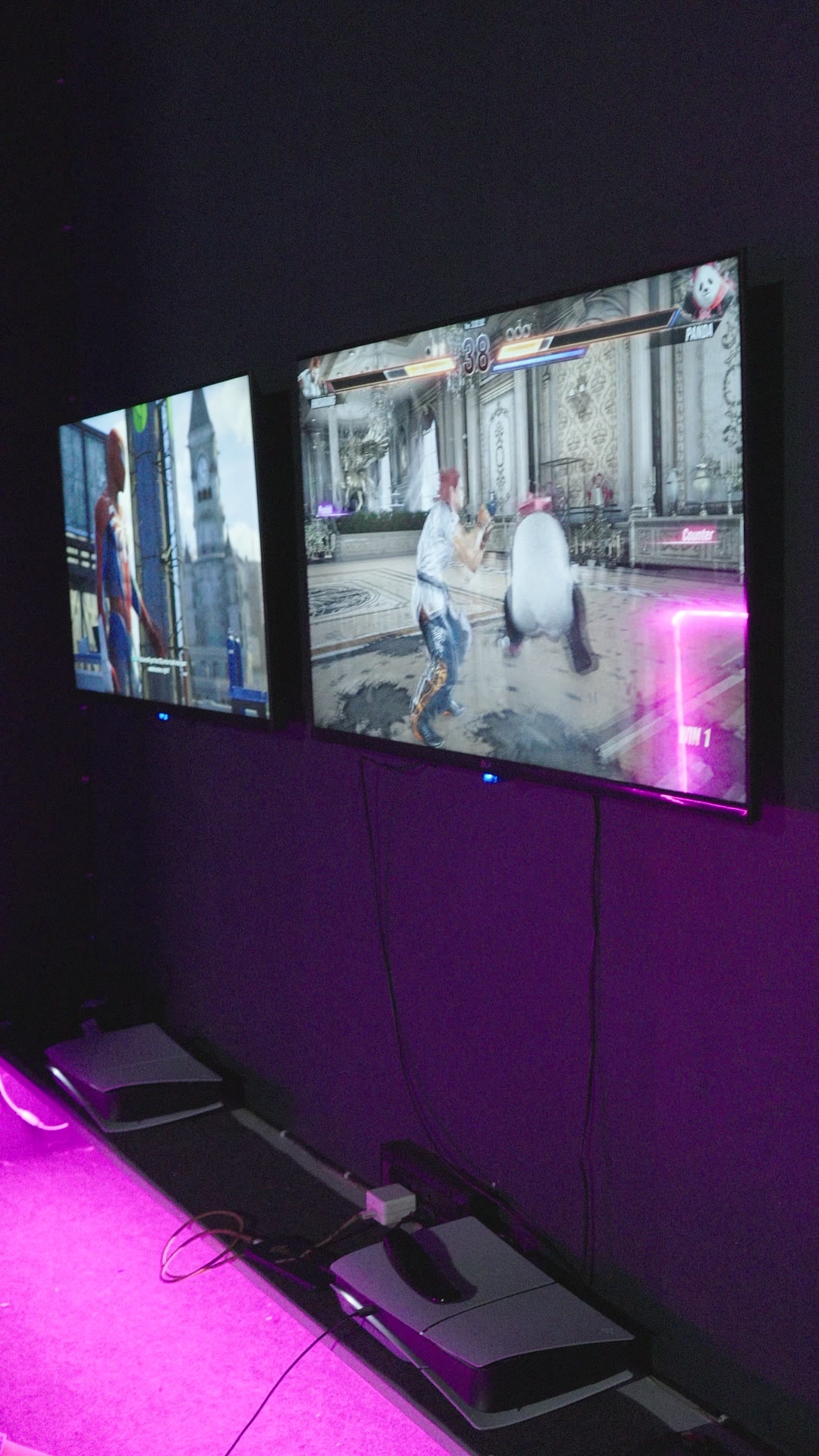 PS5 consoles on the floor below a wall-mounted TV showing a fighting game